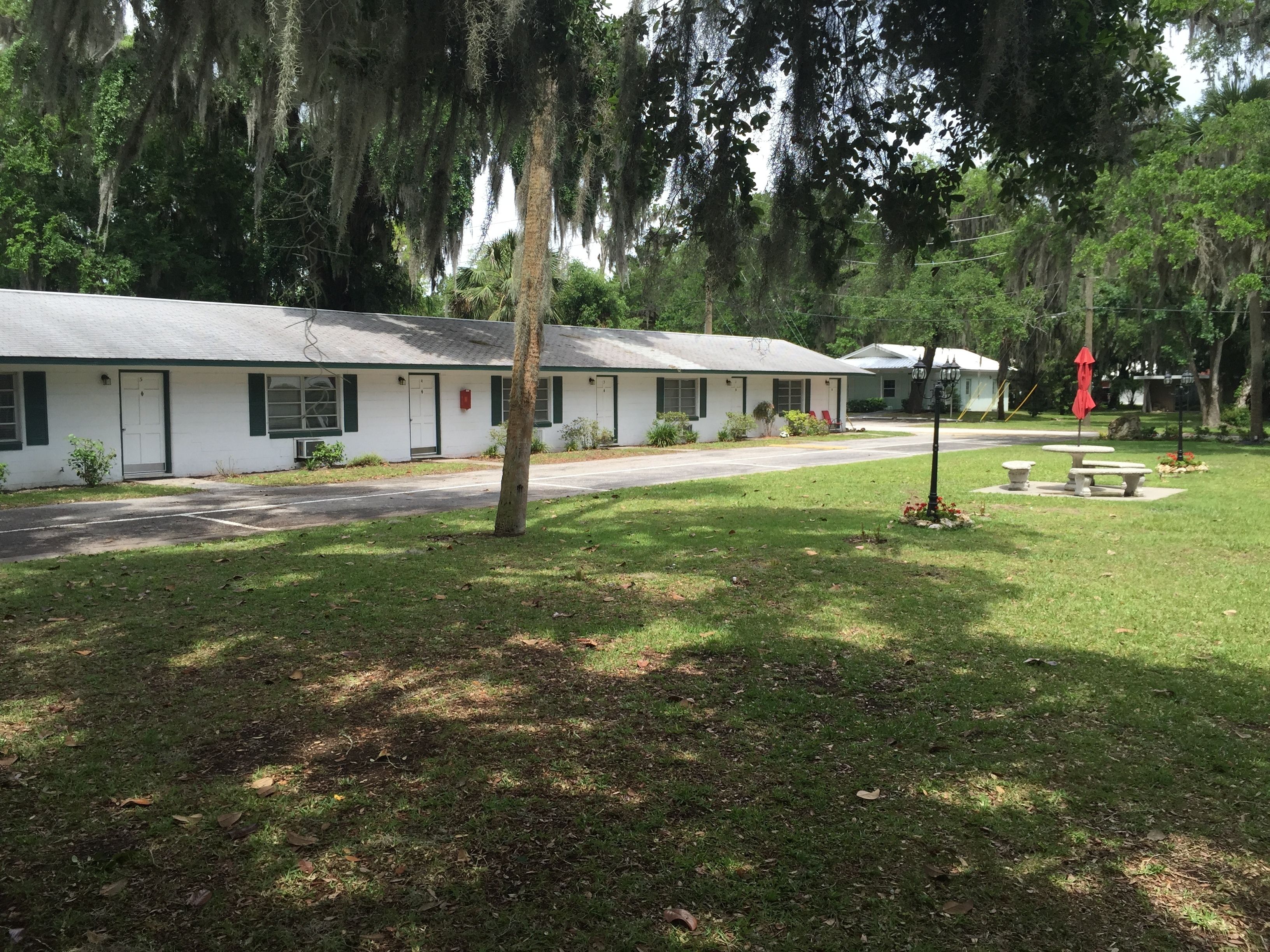 A row of white motel buildings with green shutters, surrounded by lush greenery and a well-maintained lawn.