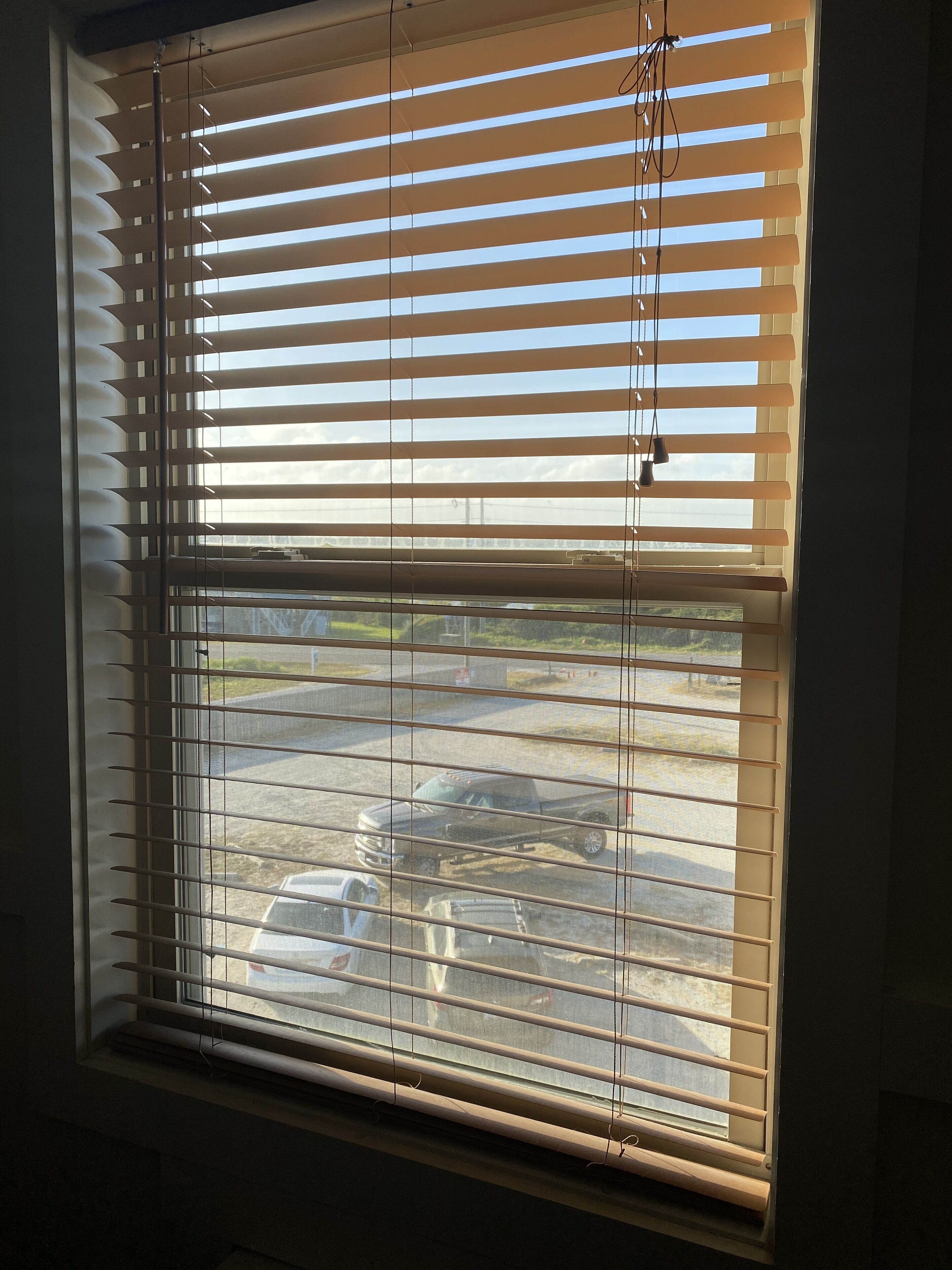A window with horizontal blinds, a view of parked cars outside, and a visible building in the background.