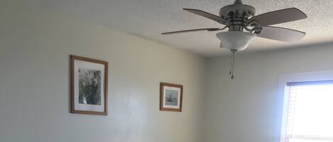A hotel room with two beds, each with a white pillow and a blue and white patterned bedspread. There is a ceiling fan, a framed picture on the wall, and a window with blinds.