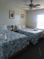 A hotel room with two beds, each with a white pillow and a blue and white patterned bedspread. There is a ceiling fan, a framed picture on the wall, and a window with blinds.
