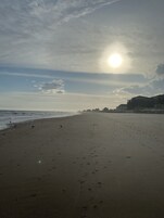 A wide sandy beach with footprints leading towards the ocean, under a partly cloudy sky with the sun visible.