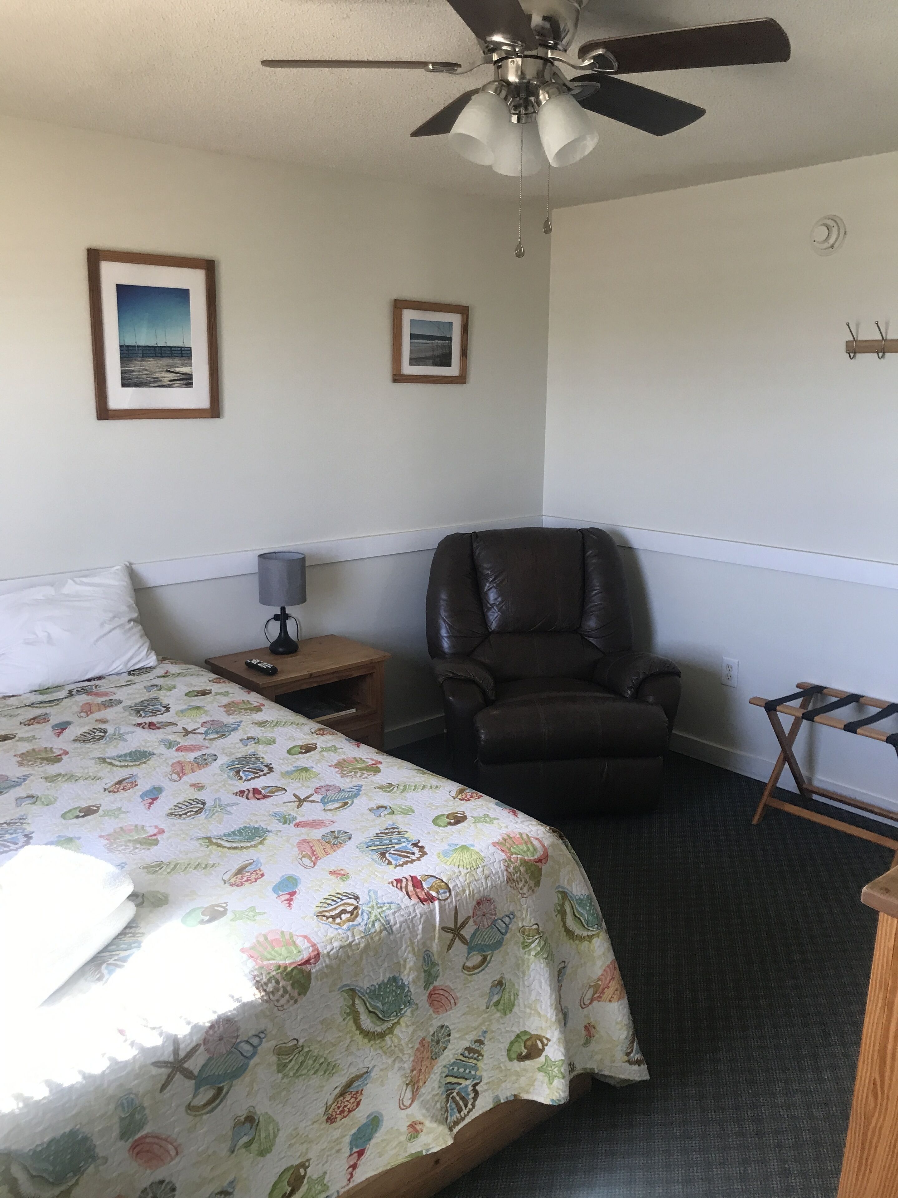 Habitación de hotel con una cama, una silla, un ventilador de techo y dos cuadros en la pared.