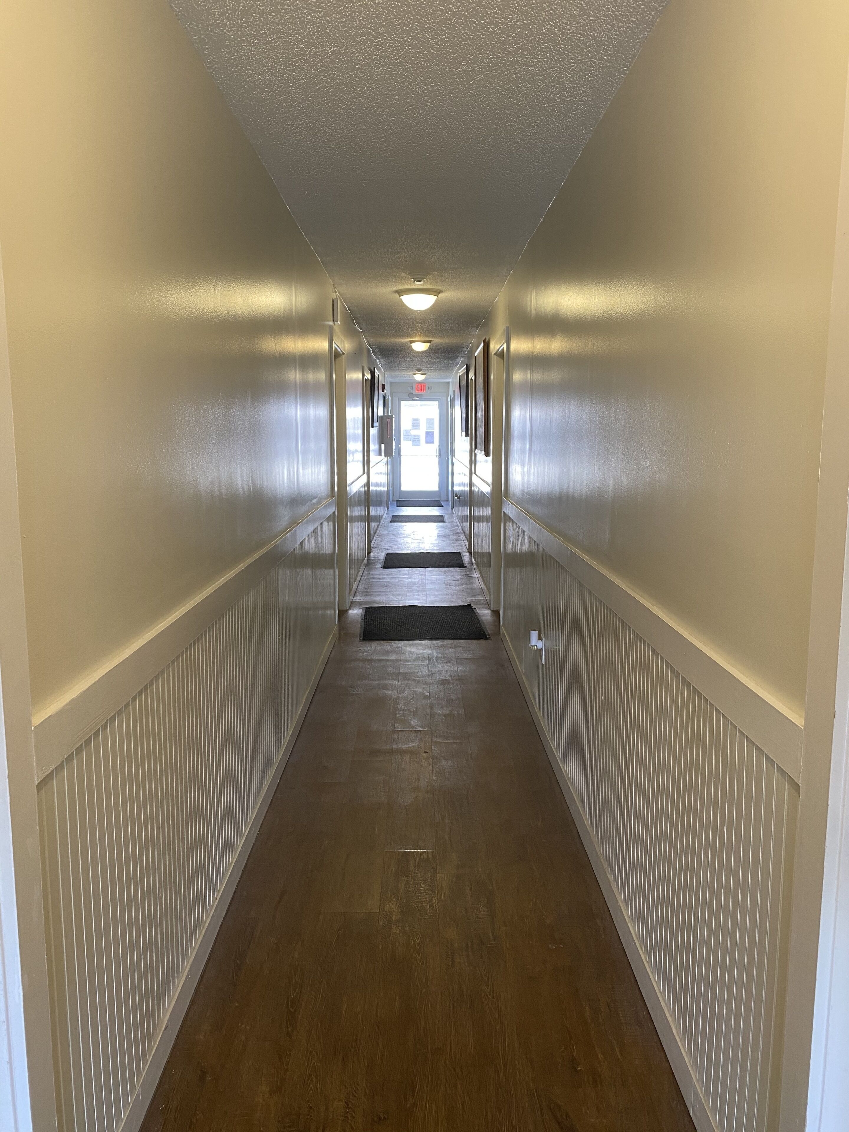 A long, narrow hallway with a wooden floor and white wainscoting on the walls.