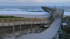 A wooden boardwalk leading to a pier extending into the ocean, with a sandy beach and grassy areas in the foreground. - Seaview Pier and Motel (Sneads Ferry)