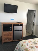 Habitación de hotel compacta con refrigerador y microondas integrados, un televisor de pantalla plana montado en la pared y una puerta que da a un pasillo.