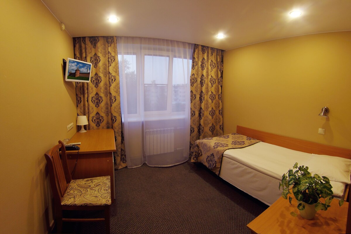 A hotel room with a bed, a desk with a chair, a small table with a plant, and a window with curtains.