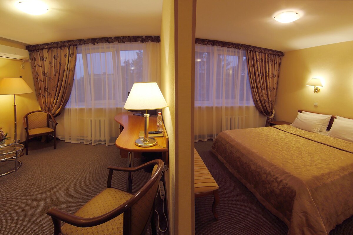 A hotel room with a bed, a desk with a lamp, a chair, and a small table.
