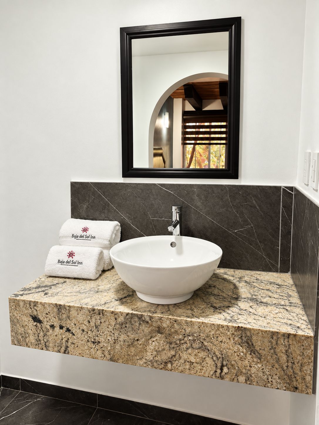 A bathroom with a white vessel sink, dark marble backsplash, and a mirror above.