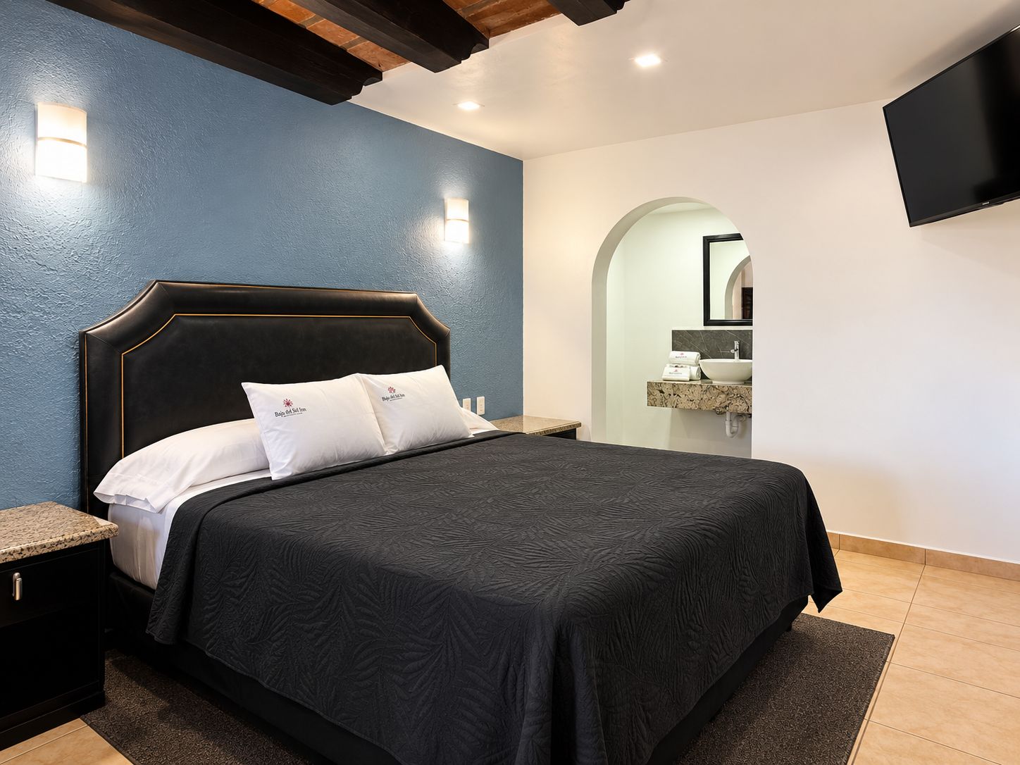 Un dormitorio moderno con una cama grande, una mesita de noche, un televisor integrado y un baño visible a través de un arco.