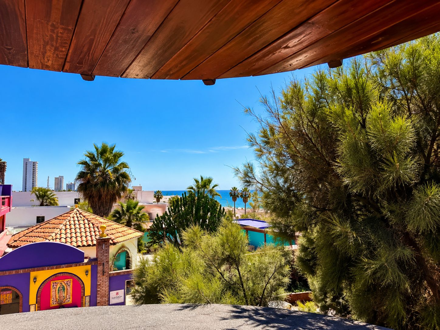 Vista costera con una pérgola de madera, un edificio colorido con tejado de baldosas y abundante vegetación.