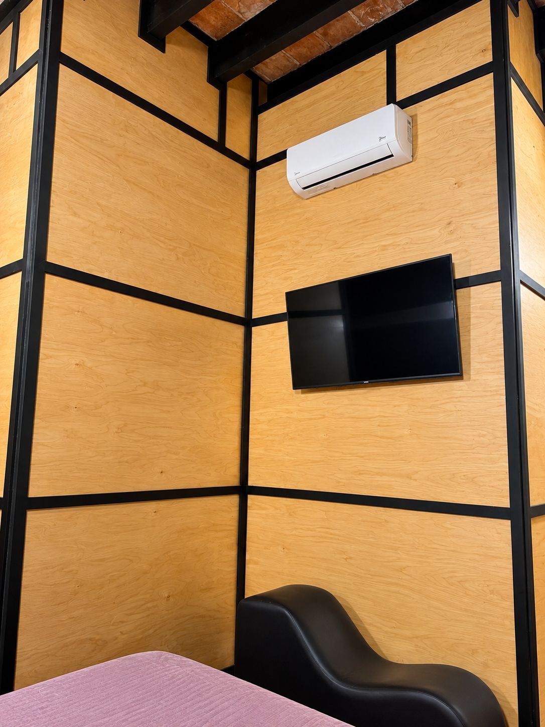 Une pièce avec un mur recouvert de panneaux de bois, un climatiseur fixé au mur, un téléviseur à écran plat et une chaise noire.