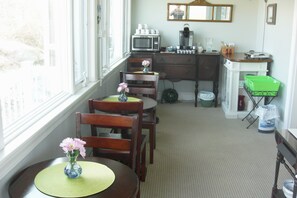 A dining area with a window, a table with a vase of flowers, and a kitchen in the background.
