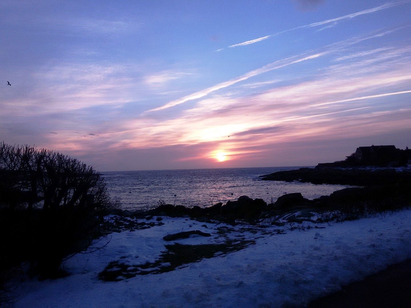 A sunset over a coastal landscape with snow-covered ground and rocky outcrops.