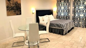 Habitación de hotel con cama, una mesita de vidrio y sillas, un cuadro en la pared y cortinas estampadas.
