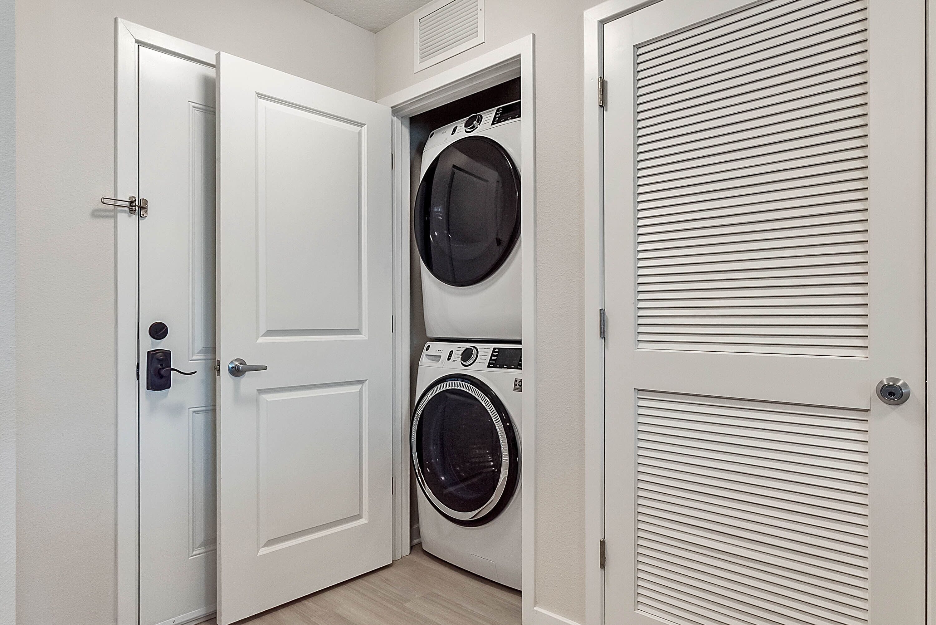 Une buanderie équipée d’un lave-linge et d’un sèche-linge à chargement frontal, d’une porte avec une poignée et d’une bouche d’aération située au-dessus du sèche-linge.
