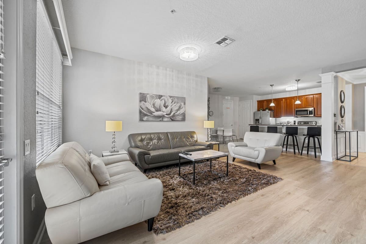Una sala de estar moderna con un sofá, sillones y una mesa de centro. Se observa una cocina con comedor al fondo.