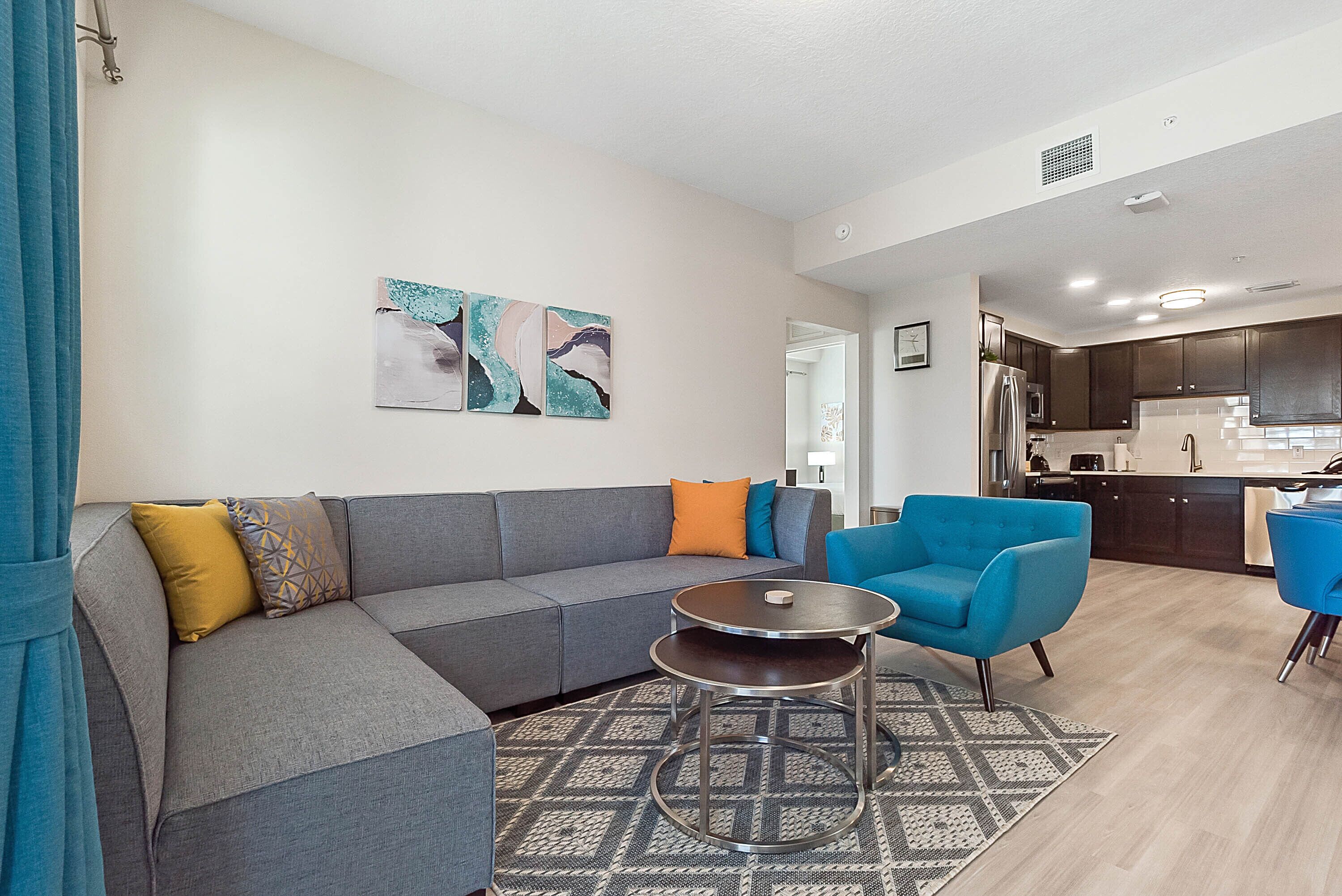A modern living room with a grey sectional sofa, a blue armchair, and a small round table. There are abstract wall art pieces above the sofa.