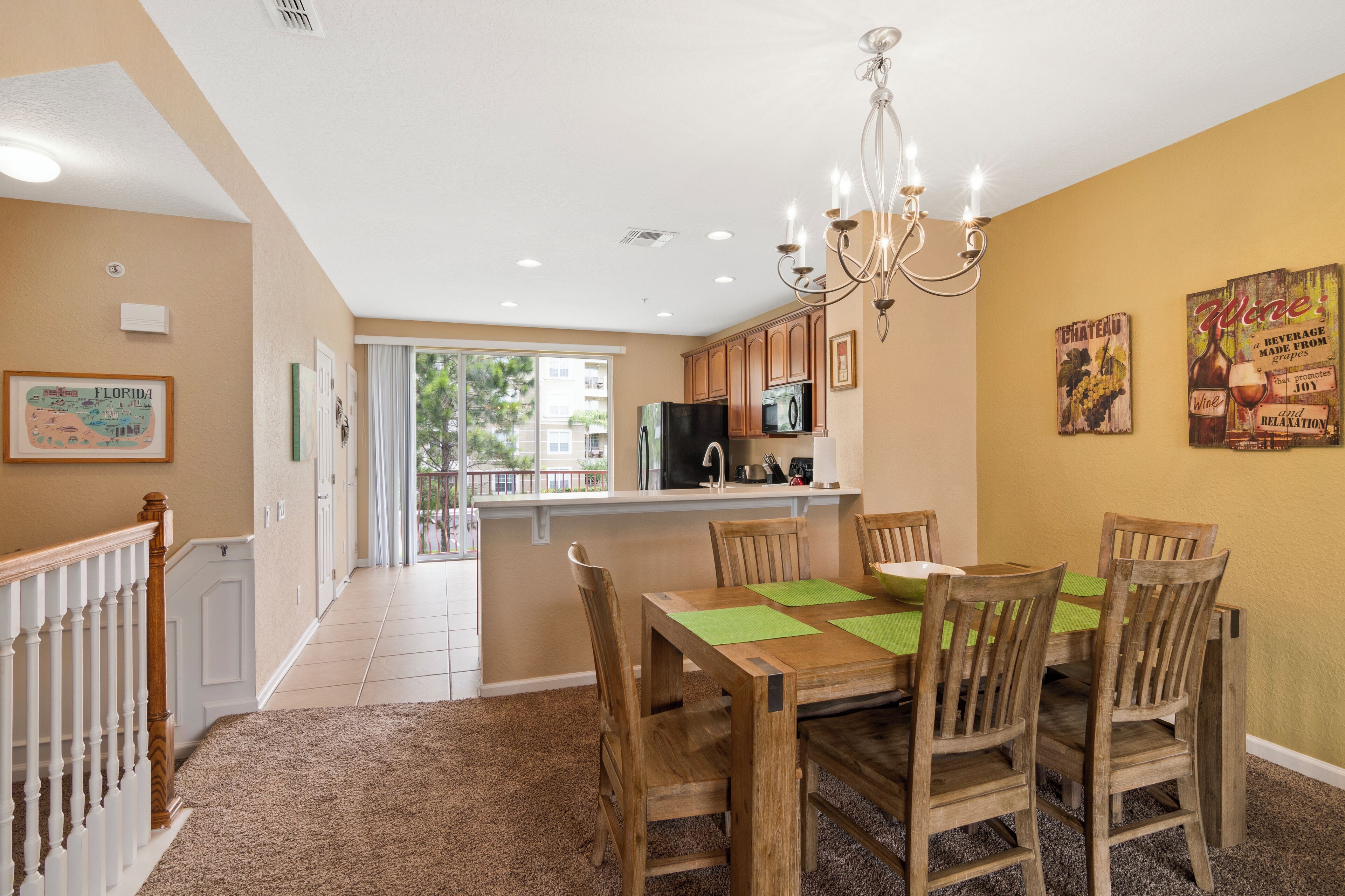 A dining area with a wooden table and chairs, a chandelier, and a kitchen in the background.