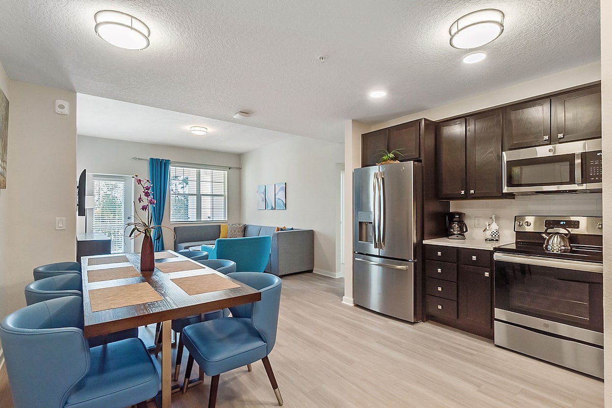 Una cocina moderna con armarios de madera oscura, electrodomésticos de acero inoxidable y una zona de comedor con sillas azules y una mesa de madera.