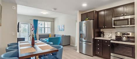 A modern kitchen with dark wooden cabinets, stainless steel appliances, and a dining area with blue chairs and a wooden table.