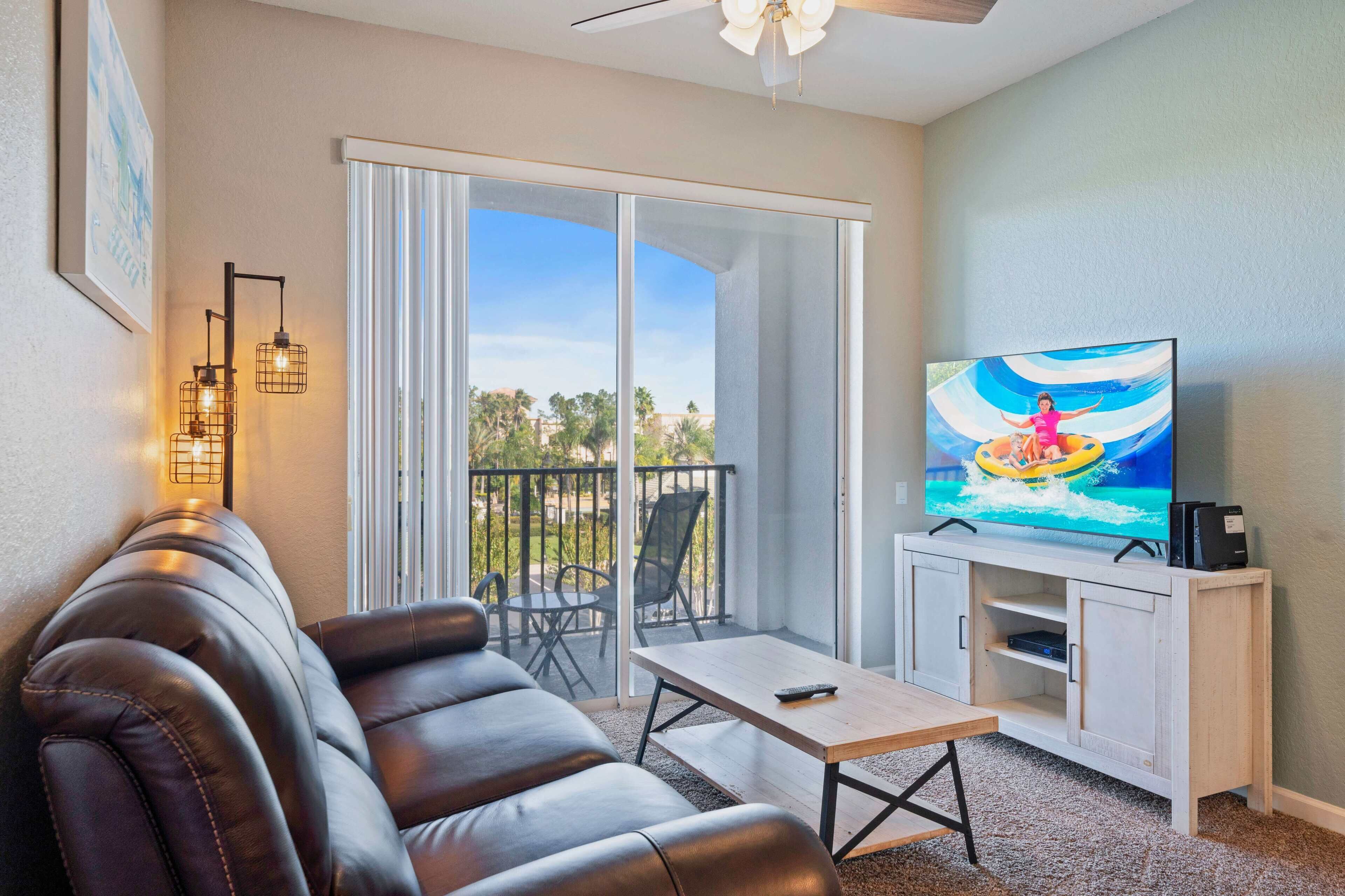 Un salón con sofá de cuero, una mesa de centro, un televisor en un mueble blanco y un balcón con vistas a palmeras.