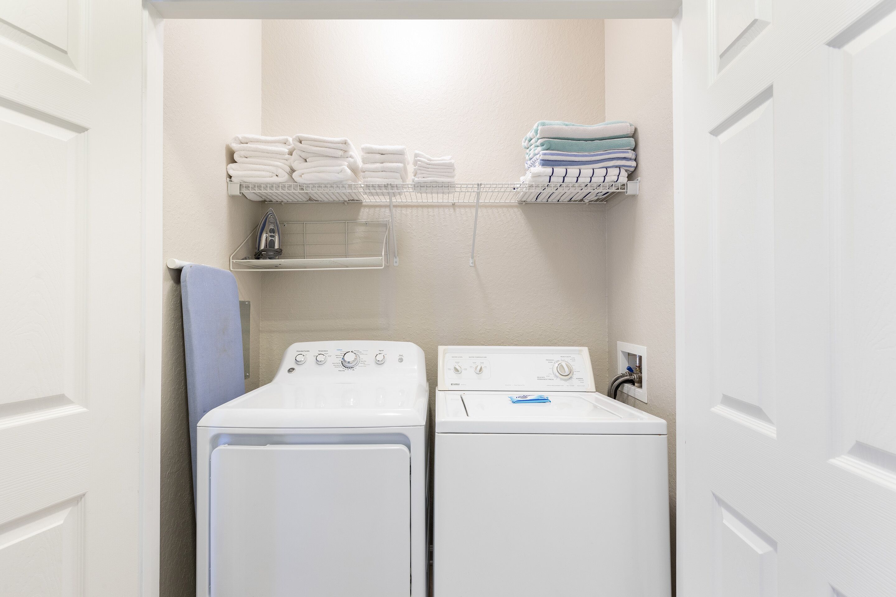 A laundry room with two white washing machines, a shelf with folded towels, and a blue shower curtain.