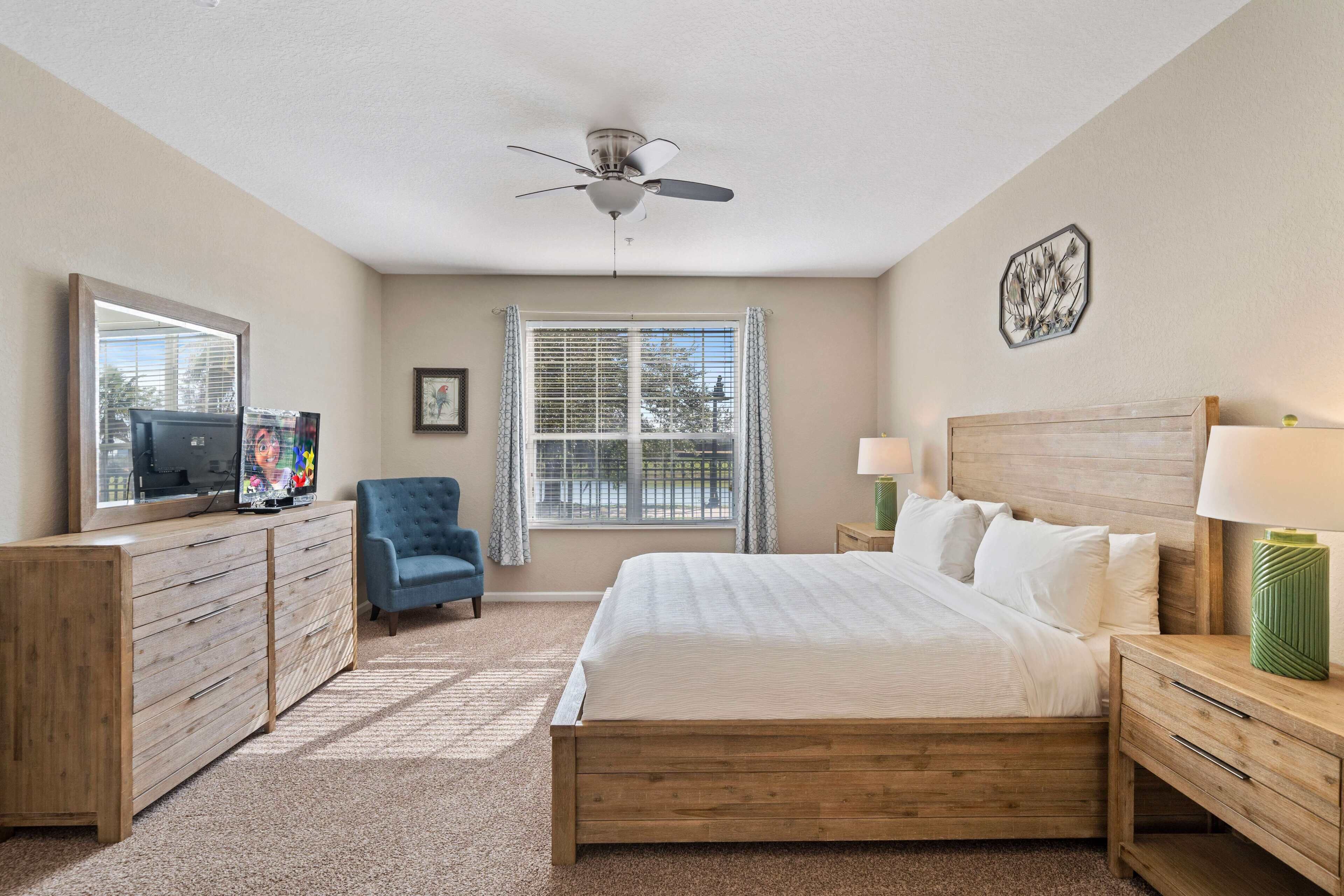 A bedroom with a wooden bed, a TV on a dresser, a chair, and a window with blinds.