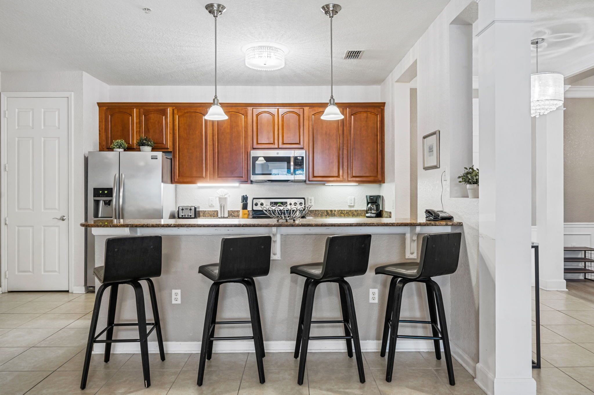 A kitchen with wooden cabinets, a stainless steel refrigerator, a microwave, and three black bar stools.