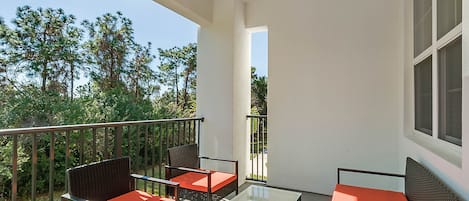 Un balcon agrémenté de meubles en osier et de tables en verre, donnant sur un jardin et des arbres.