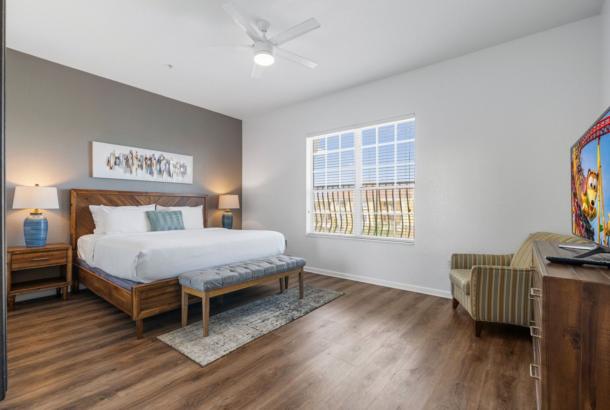 A bedroom with a bed, bedside tables, a bench, a desk, a chair, a ceiling fan, and a window with blinds.