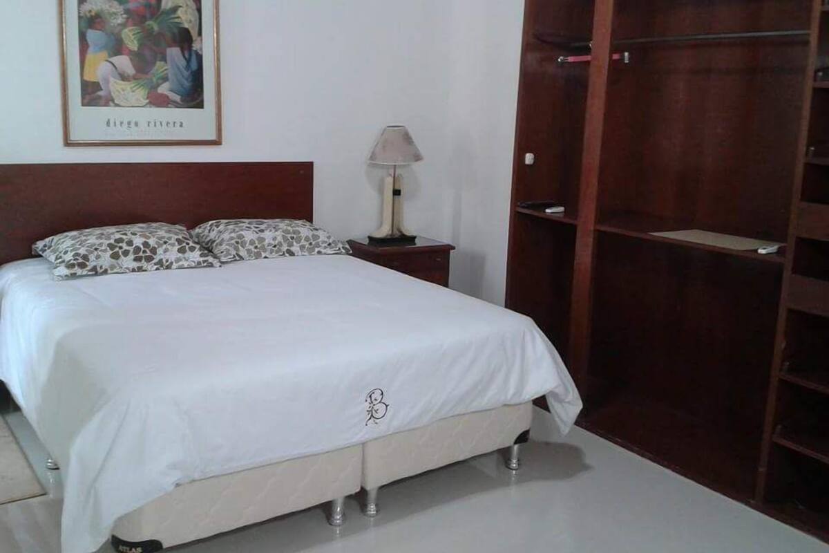 A neatly made bed with a white bedspread and patterned pillows, a wooden wardrobe, a bedside lamp, and a framed picture on the wall.