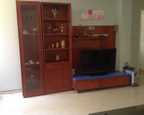 A living room with a TV on a blue platform, a wooden cabinet with glass doors, and a shelf with decorative items. - Beulah Hotel Boutique (Asunción)