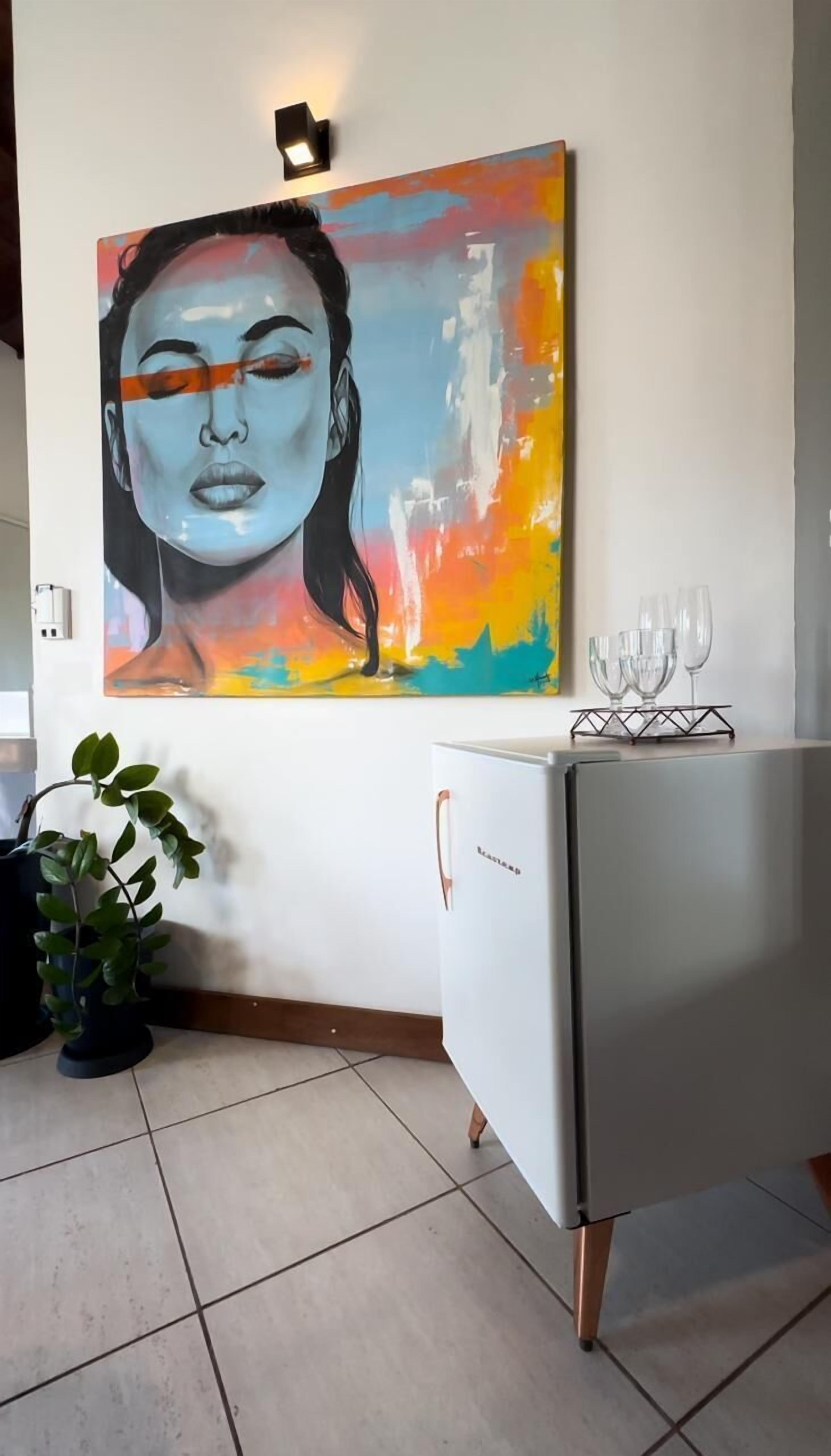 A room with a colorful abstract painting on the wall, a white cabinet with glasses on top, and a potted plant.