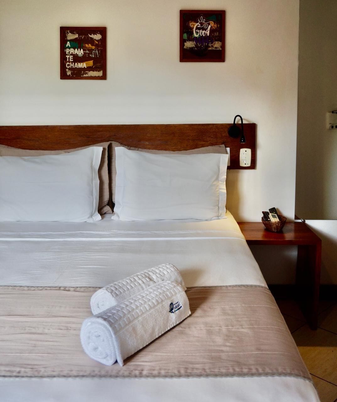 Cama bem arrumada com roupa branca e toalha enrolada, cabeceira de madeira e duas obras de arte nas paredes.