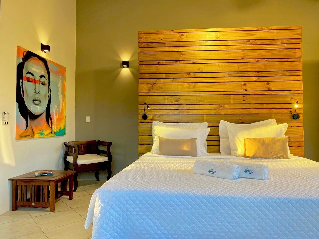 Una camera d'hotel con un poggiatesta in legno, un letto con biancheria bianca, un quadro appeso al muro, un piccolo tavolino e una sedia.