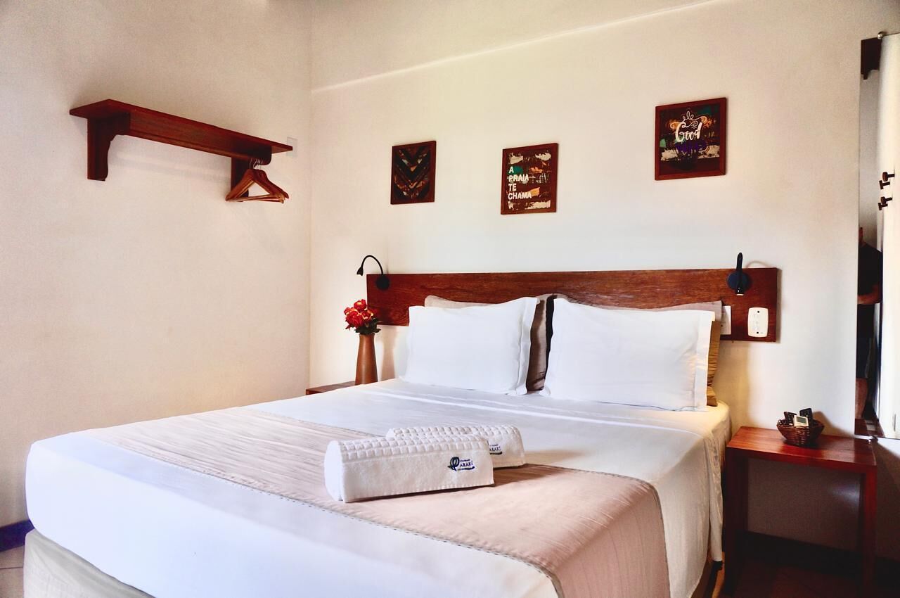 Una cama bien hecha con sábanas blancas, cabecera de madera y mesita de noche con una lámpara y un objeto decorativo pequeño.