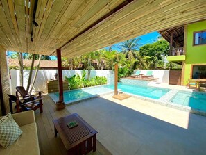 A covered outdoor area with a pool, seating, and a view of a green building. - Pousada Abaré (Itacaré)