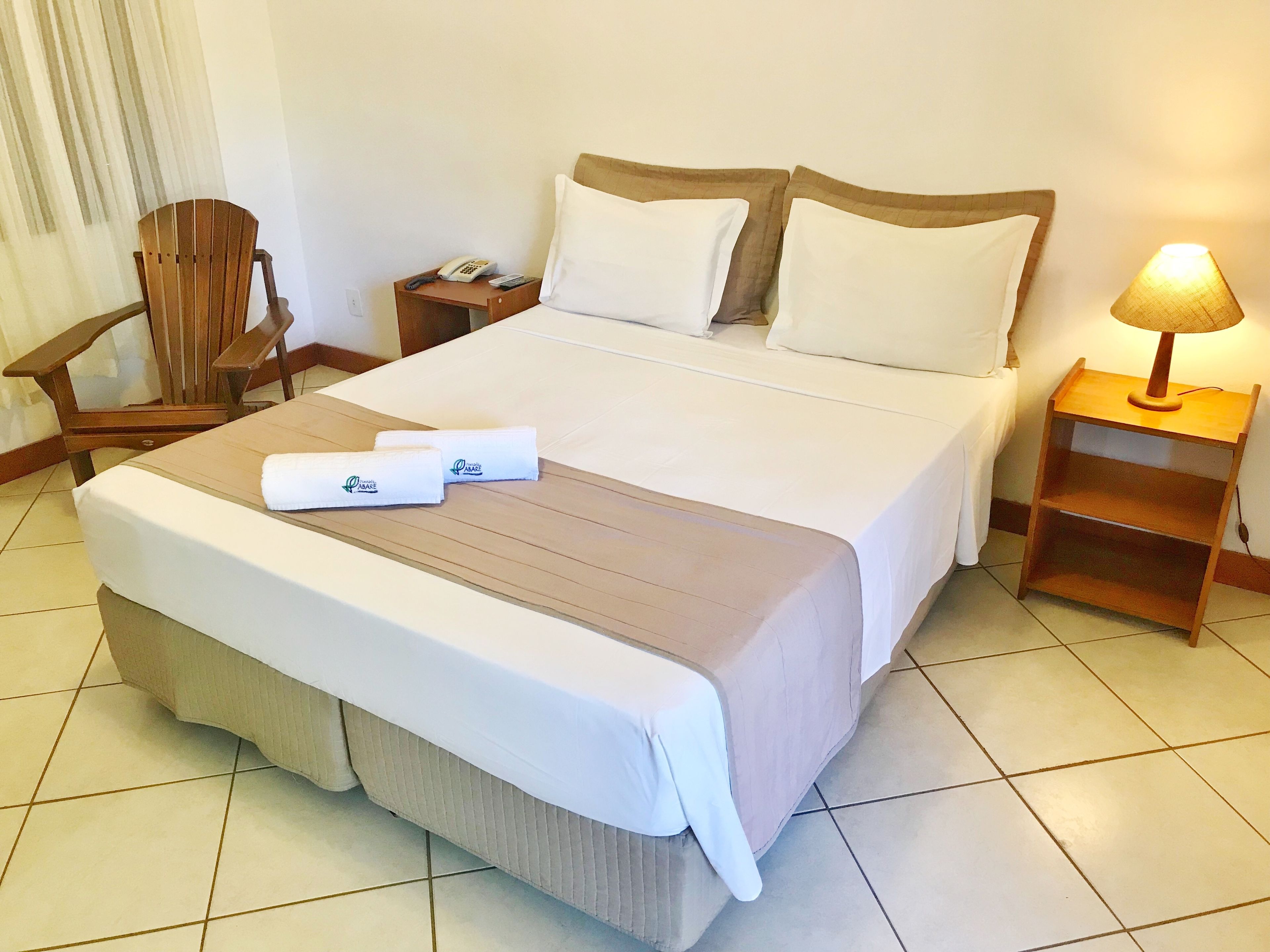 A neatly made bed with a brown bedspread, white pillows, and two rolled towels with a logo. There is a wooden chair, a nightstand with a phone and remote, and a lamp on a wooden nightstand.