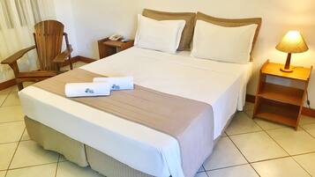 A neatly made bed with a brown bedspread, white pillows, and two rolled towels with a logo. There is a wooden chair, a nightstand with a phone and remote, and a lamp on a wooden nightstand.