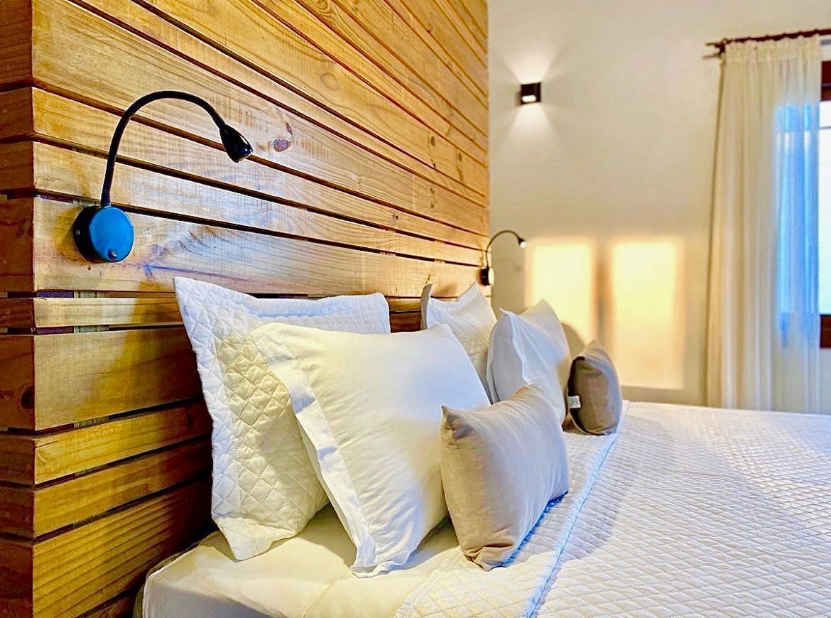 Cama bem arrumada com roupa de cama branca e várias almofadas, cabeceira de madeira, luminária de leitura fixada na parede e uma janela com cortinas.