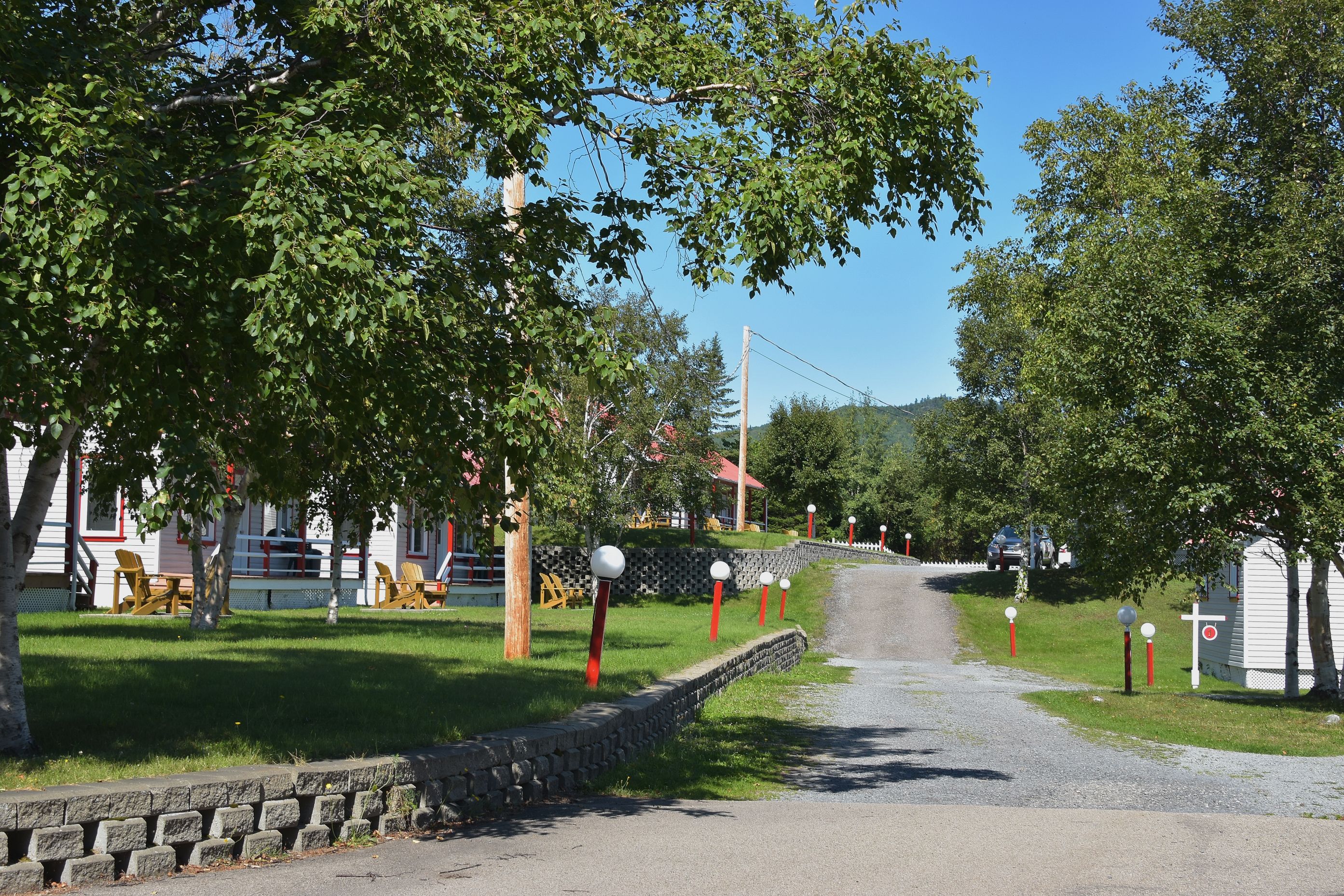 A rural road lined with trees and a stone wall. There are wooden chairs and tables in the foreground, and a white building with a red roof in the background.