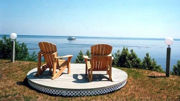 Eine runde Terrasse mit zwei Holz-Adirondack-Stühlen, die auf ein Gewässer ausgerichtet sind, umgeben von Gras und kleinen Pflanzen.