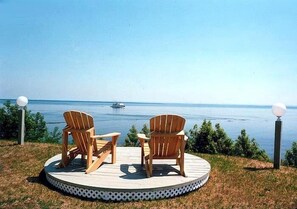 Een rond terras met twee houten Adirondack-stoelen gericht op een waterpartij, omringd door gras en kleine planten.