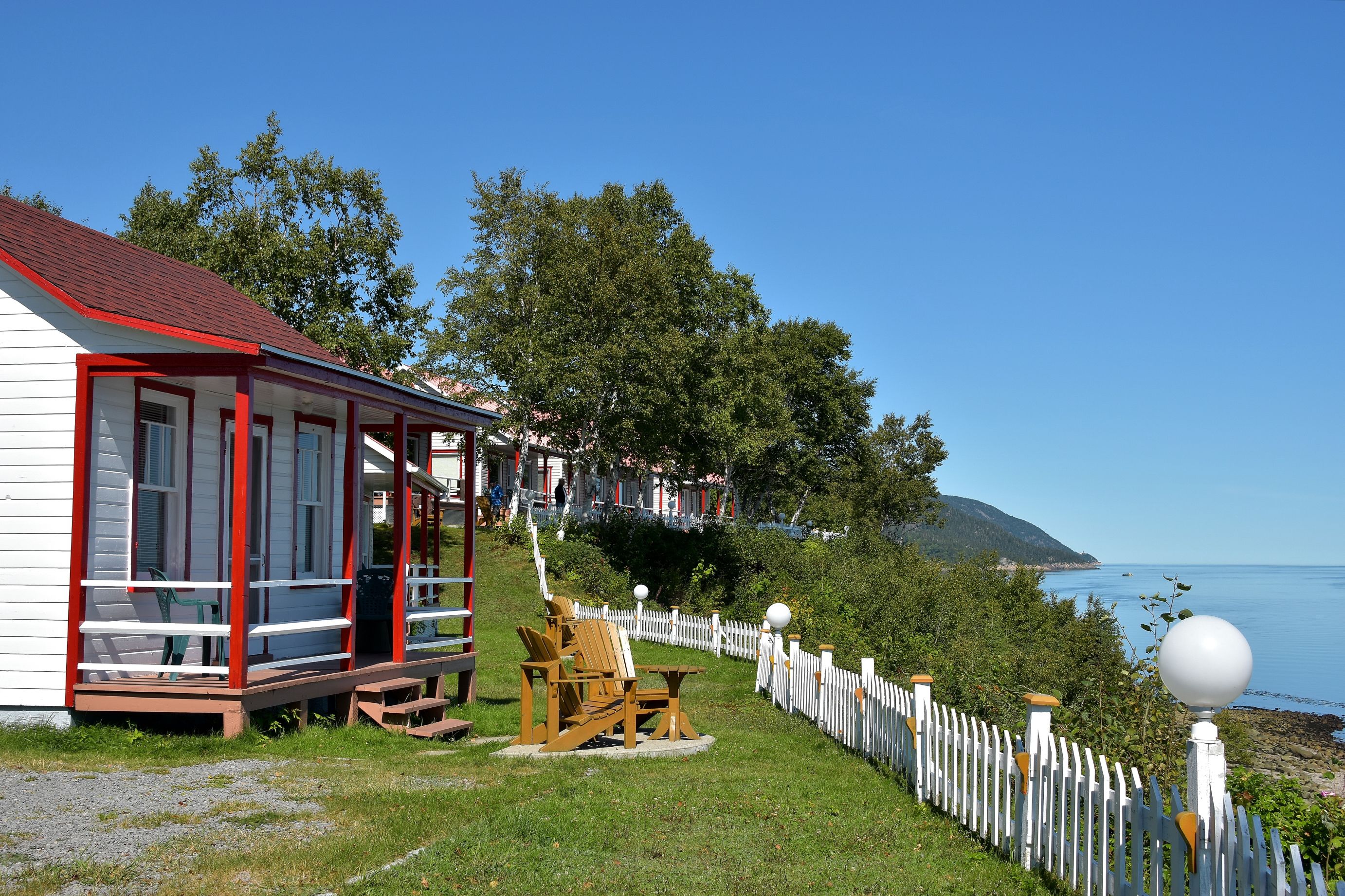 A white building with a red roof and a porch, a wooden chair and table, and a white picket fence.