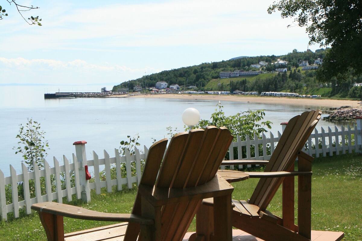 Dos sillas Adirondack de madera sobre una terraza de madera, con una cerca de piquetes blancos al fondo.