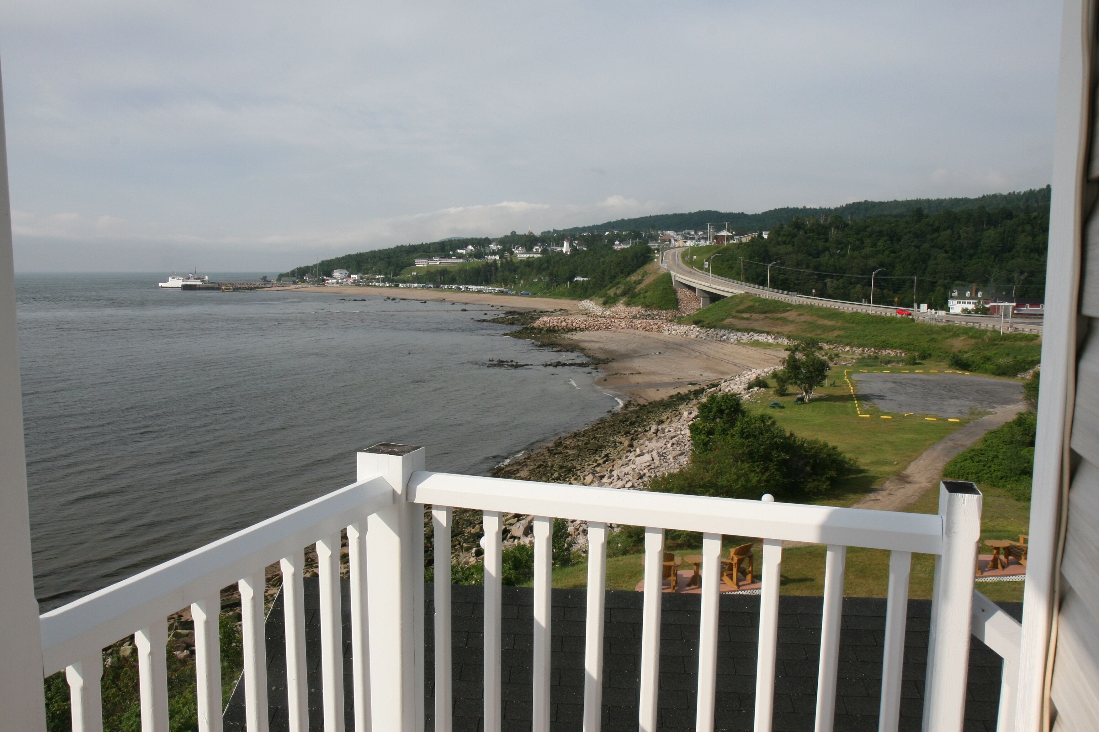A coastal view from a balcony with a railing, overlooking a beach, a road, and a hilly landscape with buildings.
