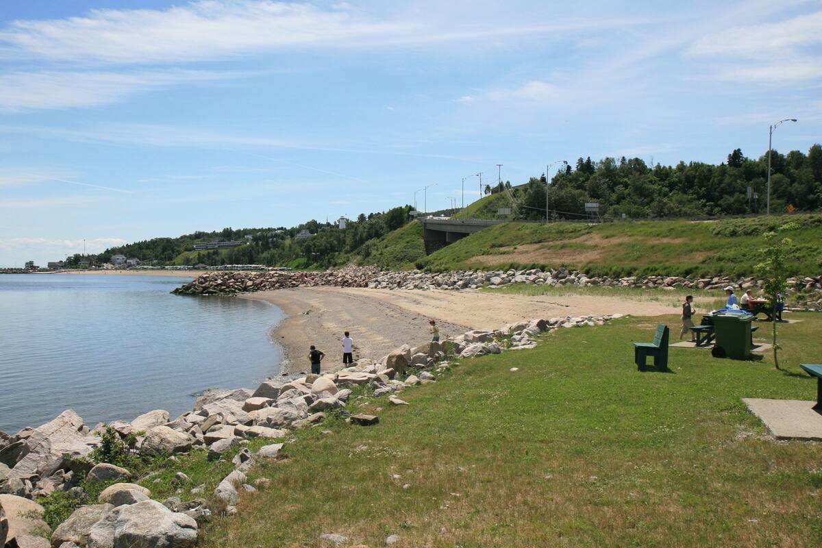 Un parque a orillas del río, con una costa rocosa, mesas de picnic y un puente al fondo.