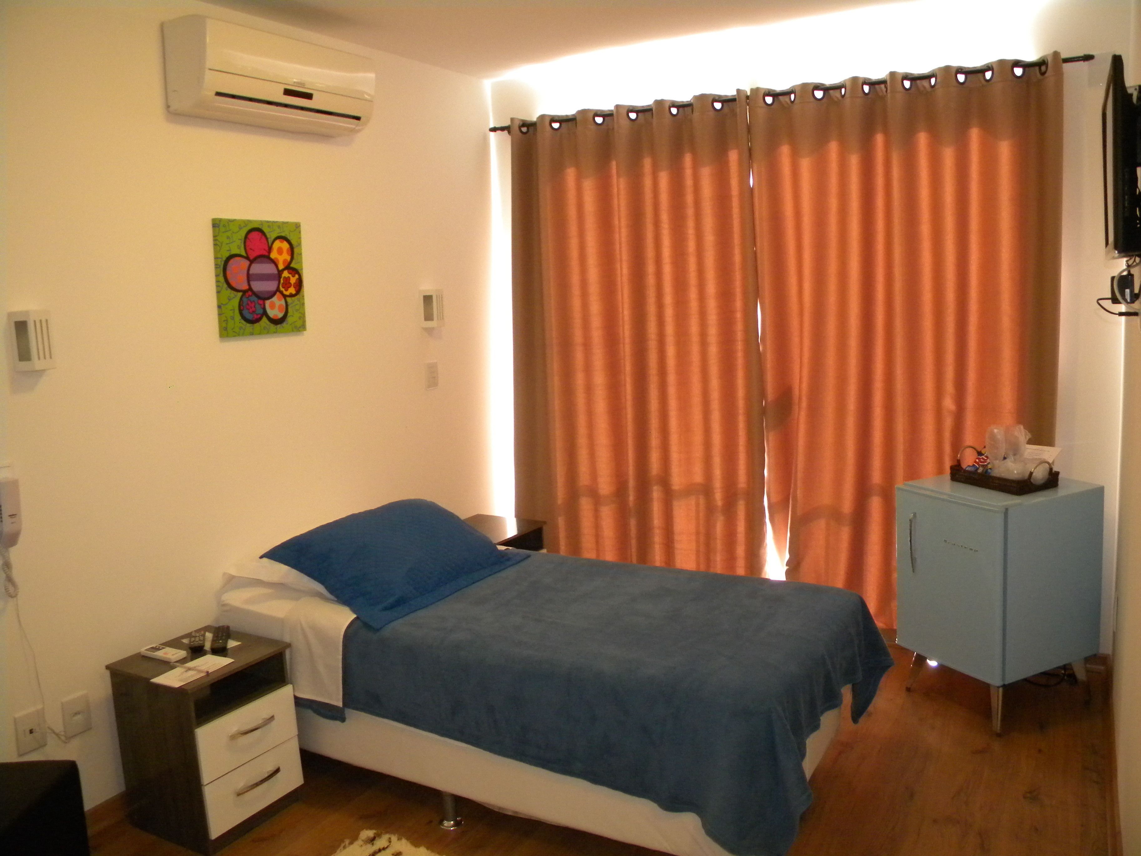 A bedroom with a bed, a nightstand, a dresser, an air conditioner, a window with curtains, and a wall-mounted TV.