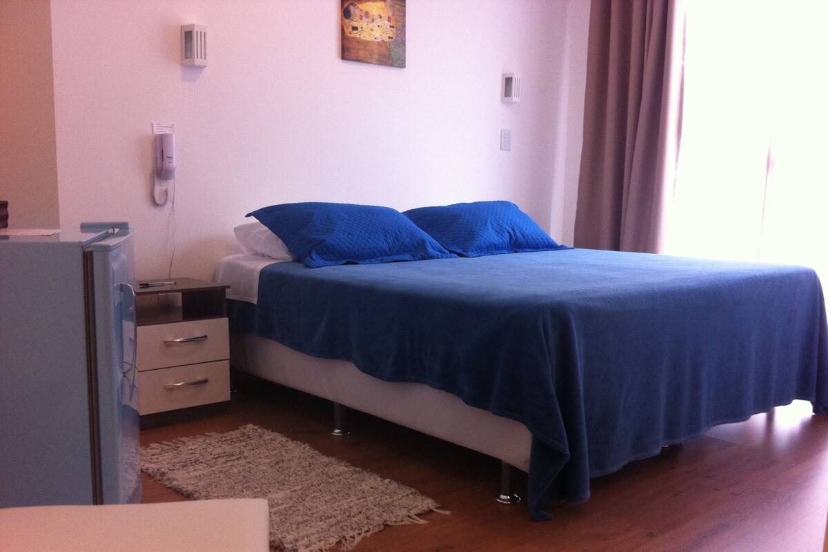 A bedroom with a bed, a desk, a chair, a refrigerator, and a painting on the wall.