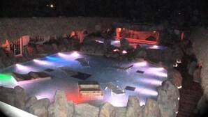 Vista nocturna de un resort con zona de piscina rodeada de rocas e iluminada por luces de colores.